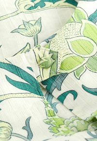 Blomster-mønstret stof med grønne og blå botaniske design på en lys beige baggrund. Inkluderer en knap og distinkte teksturer.