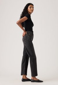 Kvinde med langt, krøllet hår iført en tætsiddende sort t-shirt, sort jeans med høj talje og sorte flade sko, stående i profil med hænderne på hoften.