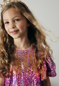 Haut à sequins brillant et multicolore avec des bordures roses, offrant une coupe décontractée et des manches courtes. Cheveux longs et ondulés, et un bandeau en forme d'étoile dorée.