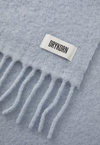 Écharpe en laine bleu clair avec une finition douce et texturée, présentant des bords à franges et une étiquette en tissu blanc avec le nom de la marque "DRYKORN."