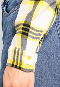 Puño de una camisa de franela a cuadros amarillos y negros con dos botones amarillos, combinada con tejido de mezclilla azul visible por debajo.