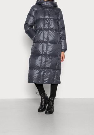 Personne portant un long manteau matelassé gris foncé avec capuche, des leggings noirs et des bottes noires à lacets, debout sur un fond uni.
