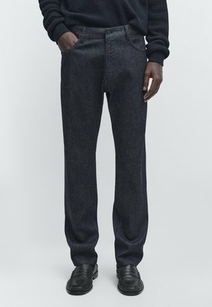 Uomo che indossa pantaloni dritti grigio scuro, mocassini neri e un maglione a coste nero, in piedi davanti a uno sfondo chiaro e semplice.