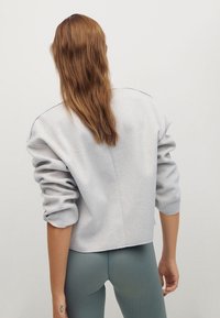 Mujer con cabello largo castaño que lleva una sudadera gris claro de gran tamaño y leggings ceñidos de canalé en color teal, de pie con la espalda hacia adelante.