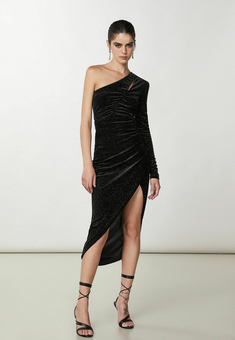 Czarna sukienka na jedno ramię z marszczonym wzorem i rozcięciem do uda, wykonana z teksturowanej tkaniny o subtelnej połysku. Czarny sandały na cienkich paskach.