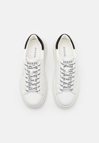 Vita sneakers med en slät läderfinish, med en svart accent vid hälen och snören som är tryckta med "GUESS" i fet svarta bokstäver.