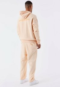 Conjunto de sudadera y pantalones de chándal color melocotón, con capucha, logotipo bordado en la parte posterior y bolsillos. Hecho de un tejido suave, con zapatillas blancas.