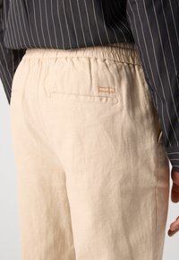 Beige Leinenhose mit elastischem Bund, ausgestattet mit einer Gesäßtasche und subtiler Textur. Gestreiftes Hemd sichtbar im oberen Bereich.