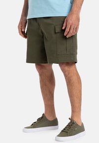 Olivengrønne cargo-shorts med sidelommer, lavet af bomuldsstof, afslappet pasform og en kant over knæet. Kombineret med grønne sneakers.