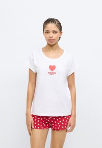 Λευκό βαμβακερό T-shirt με κοντά μανίκια, με κόκκινο γραφικό καρδιάς και τη λέξη «AMOUR». Συνδυασμένο με κόκκινα σορτς που εμφανίζουν λευκά μοτίβα καρδιάς.