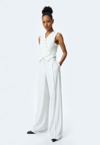 Top sin mangas blanco a rayas con botones, combinado con pantalones de pierna ancha. Ambos hechos de una tela ligera y texturizada; el modelo lleva zapatos negros de punta.