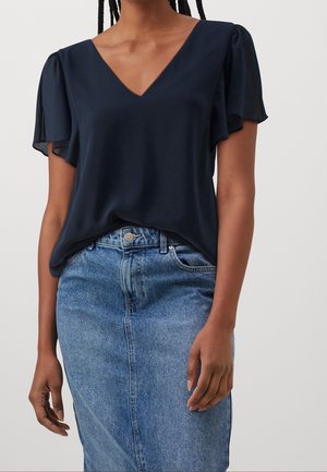 Vrouw draagt een marineblauwe V-halsblouse met fladdermouwen en een lichtblauwe high-waisted spijkerbroekrok, staand tegen een effen achtergrond.