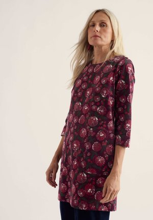 Mujer con cabello rubio largo que lleva un vestido hasta la rodilla con mangas tres cuartos y estampado floral rojo oscuro sobre fondo negro, de pie frente a una pared lisa y clara.