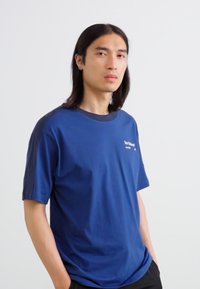 T-shirt à manches courtes bleu marine avec un col rond, présentant des accents foncés sur les épaules et un logo sur la poitrine. Tissu en coton doux.