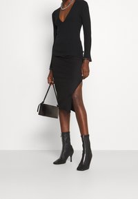 Schwarzes, geripptes Langarmshirt mit tiefem V-Ausschnitt, kombiniert mit einem schwarzen asymmetrischen Midirock. Accessoires sind eine kleine schwarze Tasche und Stiefel mit Absatz.