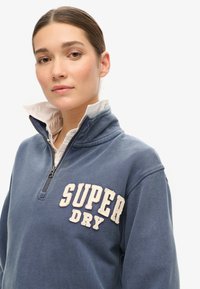Jonge vrouw met haar in een paardenstaart draagt een blauw quarter-zip sweatshirt met het logo "SUPER DRY" over een wit overhemd met kraag, op een witte achtergrond.