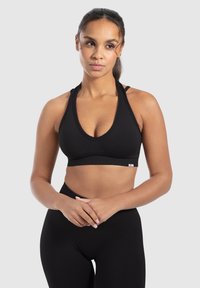 Sujeción deportiva negra con un profundo escote en V, tirantes anchos y una textura suave. Presenta una pequeña etiqueta con el logo en el dobladillo. Combinada con leggings negros.