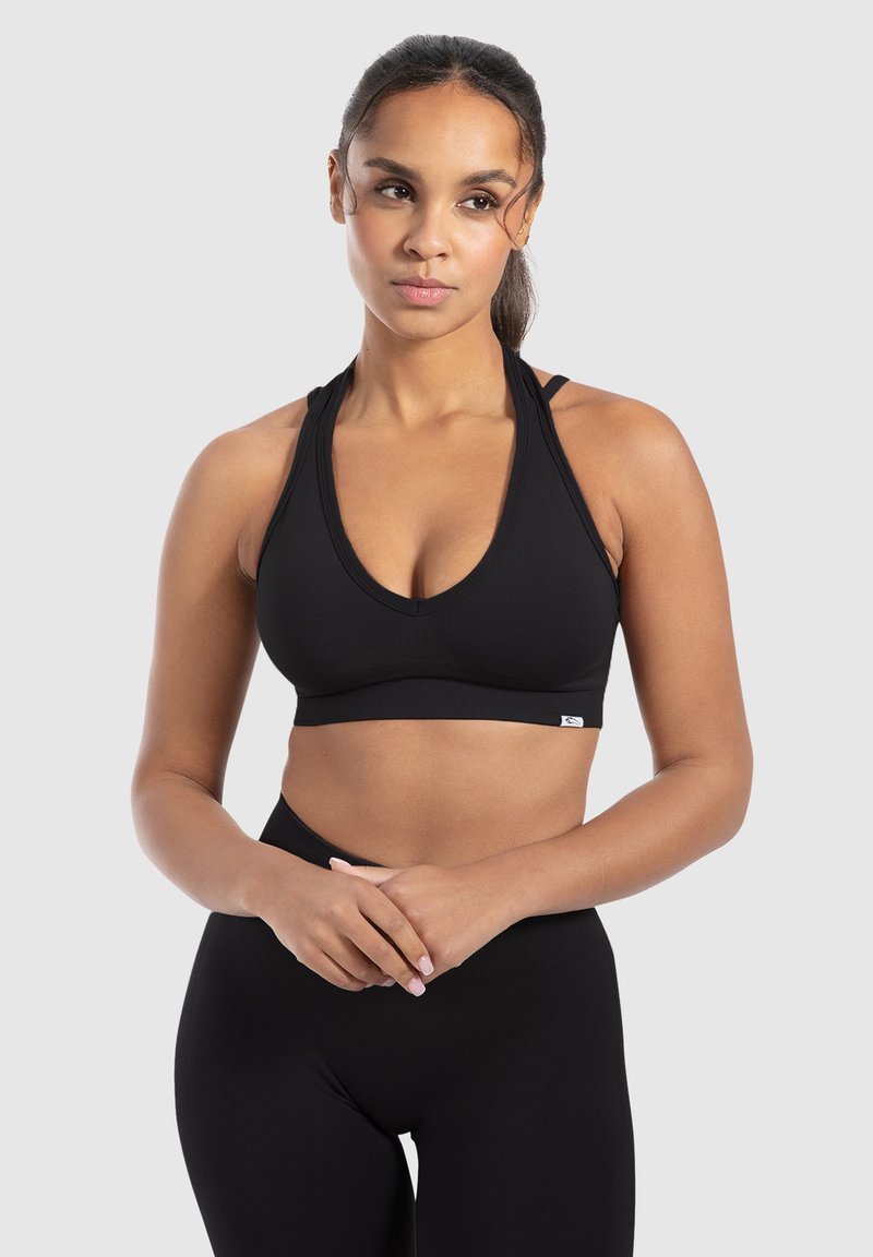 Sujeción deportiva negra con un profundo escote en V, tirantes anchos y una textura suave. Presenta una pequeña etiqueta con el logo en el dobladillo. Combinada con leggings negros.