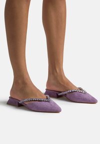 Mule in suede viola con punta affusolata, tacco blocco basso e cinturino decorato con gemme viola lungo il bordo superiore.