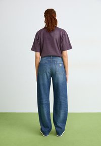Camisa de manga corta a rayas moradas y negras combinada con jeans de pierna ancha azul, que cuentan con dos bolsillos traseros y un parche de logo en la cintura.