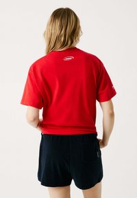Punainen puuvillainen T-paita, jossa on pyöreä kaula-aukko ja lyhyet hihat, taustalla pieni logo. Yhdistetty tummansinisiin shortseihin.
