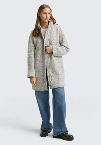 Manteau zippé gris clair texturé avec un col montant et des poches latérales, associé à un jean bleu évasé et des chaussures noires à semelle épaisse.