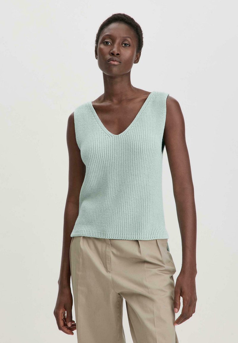 Lichtgroene mouwloze gebreide top met een V-hals, ribbelstructuur en een rechte zoom, gecombineerd met beige broek.