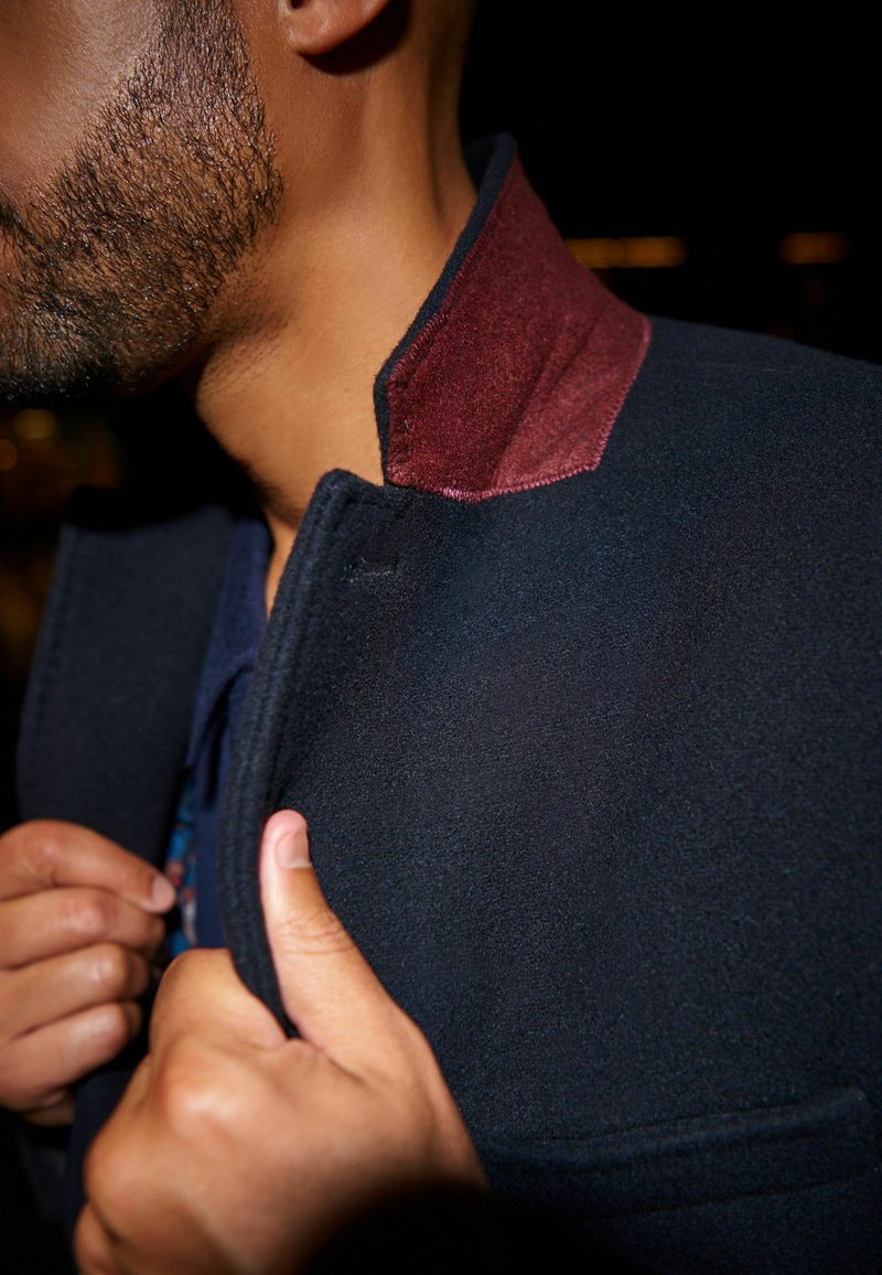 Abrigo de lana azul marino con un cuello de ante burdeos en contraste, que presenta una textura suave y un diseño estilizado. Primer plano del cuello y de la parte delantera abrochada.