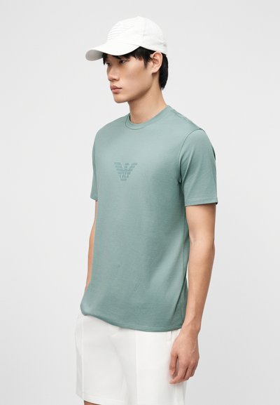 Άνδρας που φοράει ανοιχτό πράσινο κοντομάνικο πουκάμισο με λογότυπο, λευκά σορτς και λευκό καπέλο, στέκεται πλάγια μπροστά σε απλό φόντο.