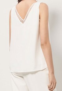 White sleeveless top featuring a V-back with lace detailing, smooth texture, and a relaxed fit, ideal for casual wear.