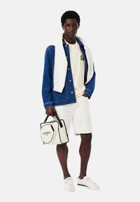 Giacca di denim, t-shirt crema, pantaloni shorts bianchi e sneaker bianche. Porta una borsa rettangolare Lacoste crema con dettagli neri.