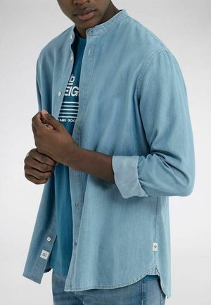 Hombre con camisa vaquera azul claro con las mangas remangadas y camiseta azul con diseño gráfico debajo, ajustándose los botones de la camisa.