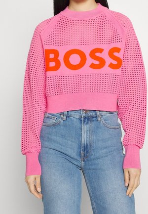 Kobieta ubrana w różowy dziergany sweter z wzorem kratki i wyraźnym czerwonym napisem "BOSS", zestawiony z wysokim stanem niebieskich dżinsów.