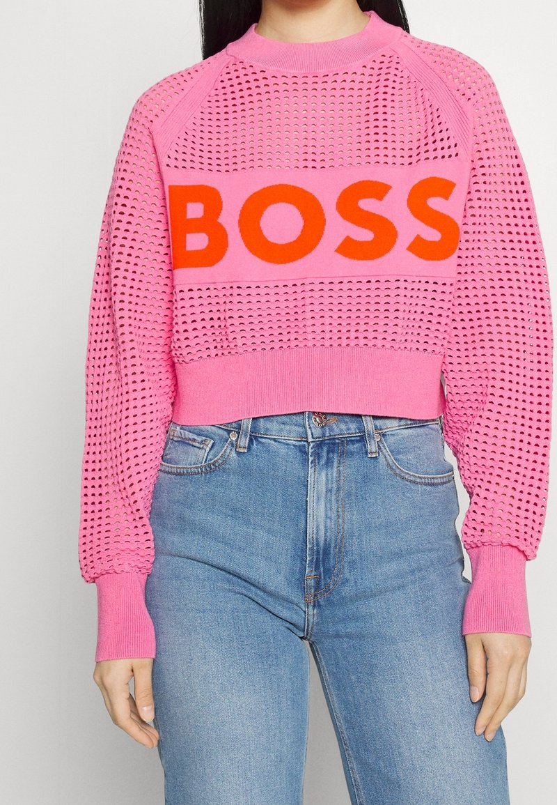 Femme portant un pull rose en maille avec un motif en treillis et un texte rouge vif "BOSS", associé à un jean bleu taille haute.
