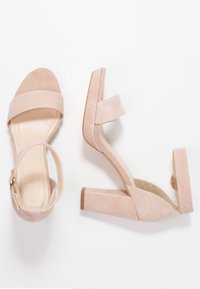 Pár béžových semišových sandálů na blokovém podpatku s popruhy kolem kotníku na bílém pozadí, jedna bota leží plocho a druhá je na boku.