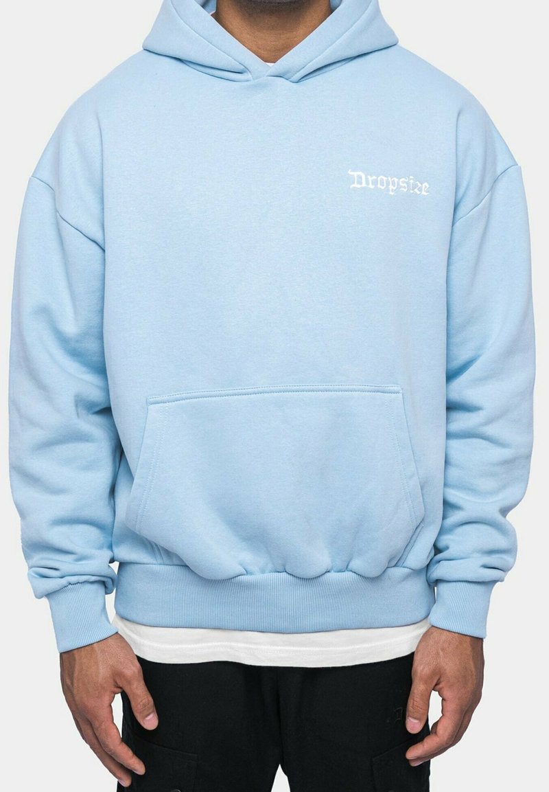 Personne portant un sweat à capuche bleu clair avec une poche avant et un texte blanc "Dropsize" sur la partie supérieure gauche de la poitrine, assorti à un pantalon noir et un maillot blanc en dessous.