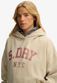 Beżowa bluza z kapturem z przednią kieszenią kangur, z dużym czerwonym tekstem "S.DAY" i "N.Y.C." Wydrukowana na miękkiej tkaninie przypominającej polar.