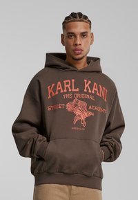 Sudadera marrón de corte oversize, con una impresión gráfica naranja de un águila y el texto "KARL KANI THE ORIGINAL STREET ACADEMY."