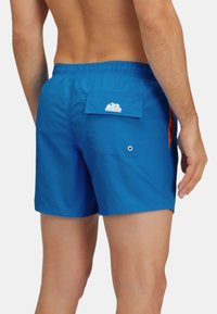 Pantaloni da bagno blu con vita elastica, tasca posteriore con logo a nuvola bianca e accento arancione sul lato. Tessuto dalla texture liscia.