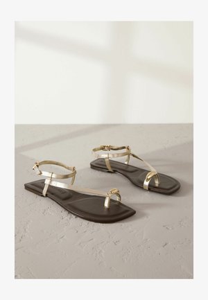Sandale plate cu talpă maro închis și bretele metalice aurii. Designul include un separator pentru degetul mare și o curea la gleznă cu cataramă ajustabilă.