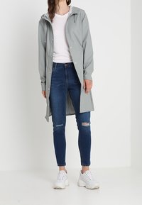 Jasnoszary płaszcz przeciwdeszczowy z kapturem, zapinany na zatrzaski, w połączeniu z ciemnoniebieskimi podartymi dżinsami i białymi trampkami. Gładka faktura, długość do kolan.