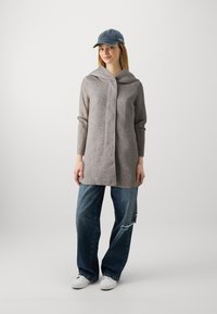 Lys grå hettegenser med tekstur, kombinert med slitte blåjeans og hvite joggesko. Tilbehør inkluderer en blå caps.