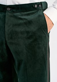 Close-up of dark green velvet trousers with dual black buttons at the waistband and a hand tucked into the right pocket.