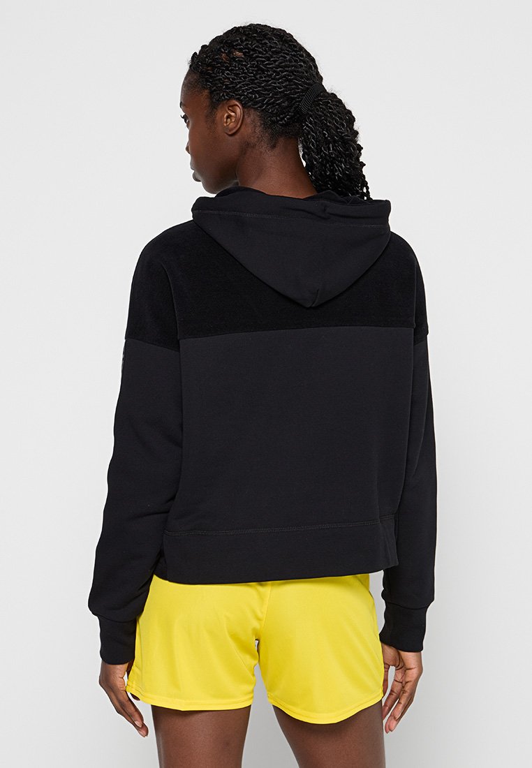 Schwarzer Kapuzenpullover mit strukturiertem oberen Schulterbereich, cropped Design und weichem Stoff. Getragen mit leuchtend gelben Shorts, die einen Farbkontrast zeigen.