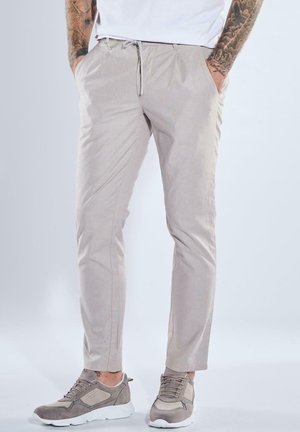 Man wearing beige slim-fit pants and gray sneakers, standing with hands in pockets against plain background.