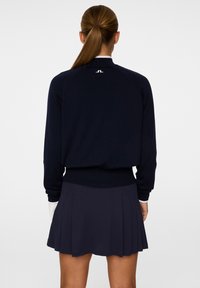 Laivastonsininen neulepaita, jossa on ribbaus hihansuissa ja helmassa, takana valkoinen logo. Yhdistetty laivastonsiniseen pliseerattuun hameeseen, luoden yhteensopivan ilmeen.