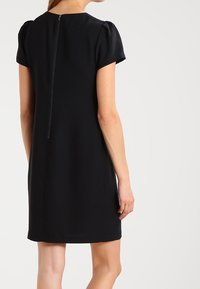 Robe noire avec des manches courtes bouffantes, un col rond et une fermeture éclair au dos. Tissu lisse, longueur genoux avec une légère forme évasée.