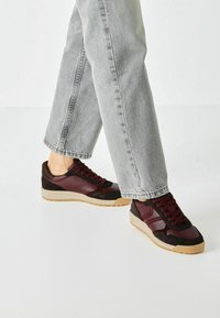 Burgundy and black sneakers with fabric and leather upper, rubber sole, and red laces. Paired with light grey wide-leg jeans.