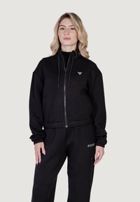 Juodas zip-up džemperis pagamintas iš minkštos medžiagos, su pritaikytu juosmeniu, reguliuojamomis raišteliais ir maža logo ant krūtinės.