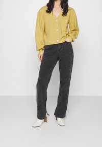 Žena v žlutém knoflíkovém kardiganu, černých rovných džínách s bočními rozparky a bílých kotníkových botách na podpatku, stojící s rukou v kapse.